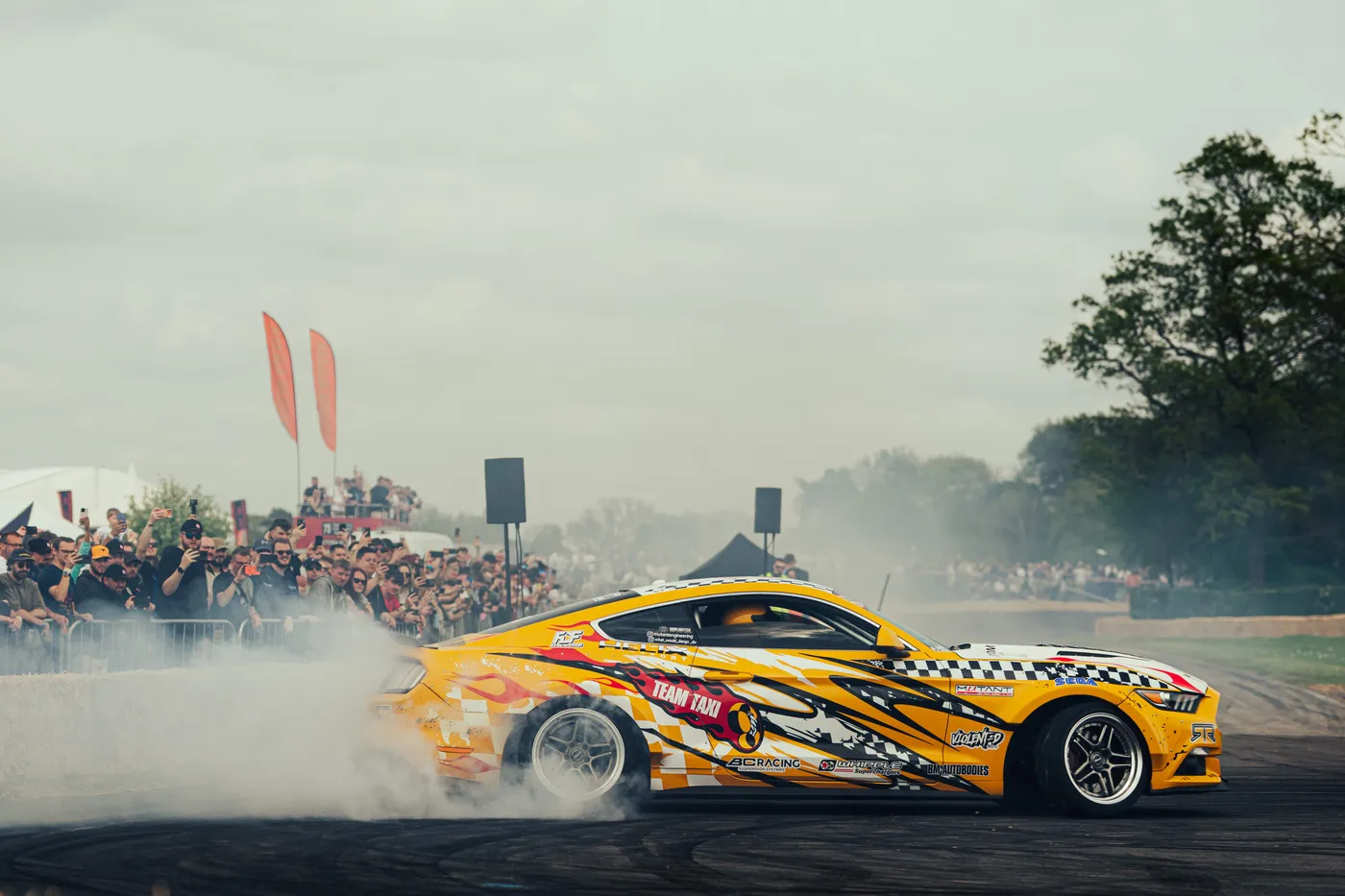 A liveried Ford Mustang drifting in a cloud of tyre smoke past a packed crowd line at ShedFest